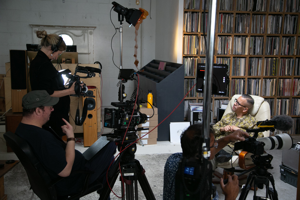 Troi lee sitting in an arm chair with filming equipment in shot. You can see the film crew